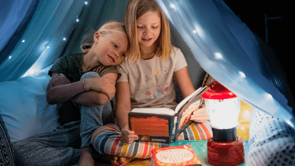 Two kids read a book out loud together in a blanket fort with fairy lights.