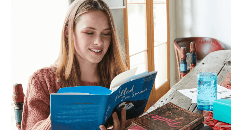 A teenage girl sits at the kitchen table reading a book.