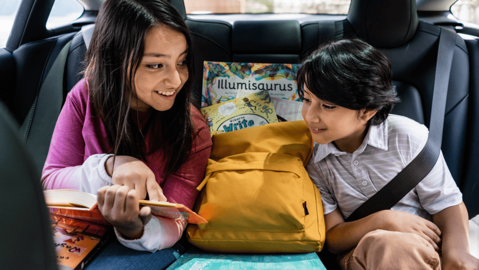 Sister and brother look at a book in the back seat of a car. A yellow book bag and Literati box are between them.