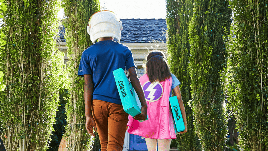 A boy in an astronaut helmet carries a Literati box up the drive, with his sister dressed as a super hero