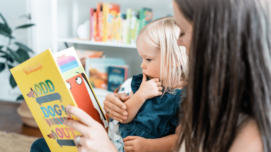 A mother reads a book aloud to her toddler who looks pensive.
