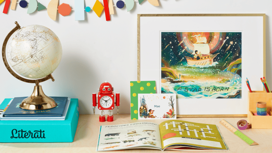 A small red robot sits on a desk near a Literati Box, globe, open book, framed picture and school supplies.