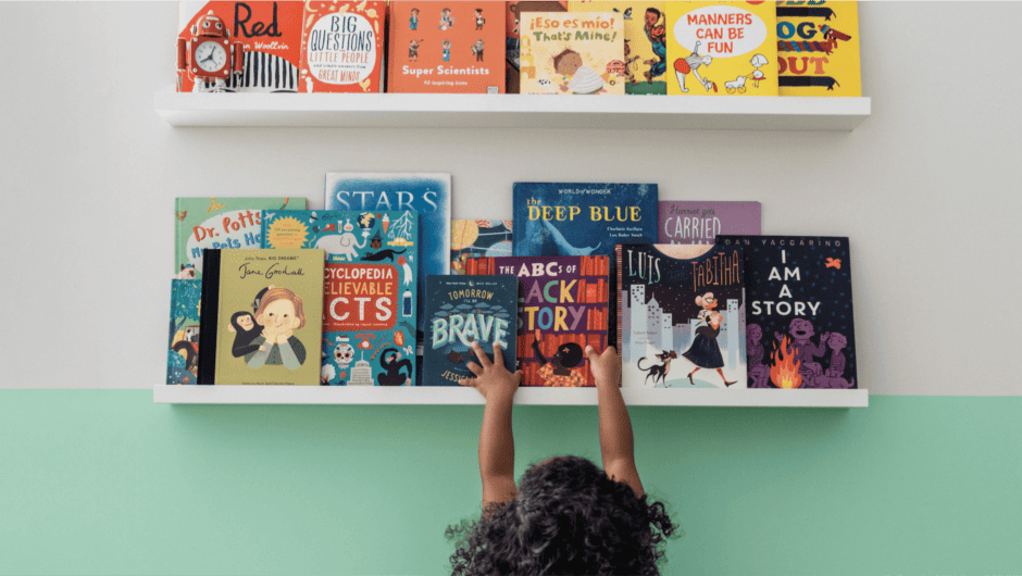 A young girl reaches for a book on a shelf. The collection of books in the order of a rainbow.