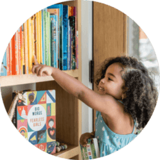A young girl points a book spine on a shelf. The books are arrange in rainbow order.