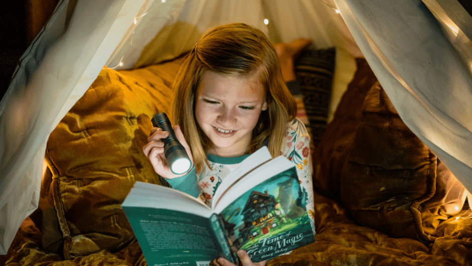 Young girl reads a book in the dark with a flashlight in a bed fort.