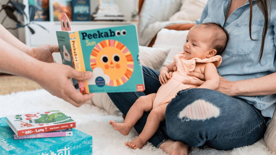 A very young baby smiles while being held by her mom, someone else holds a board book for her to see
