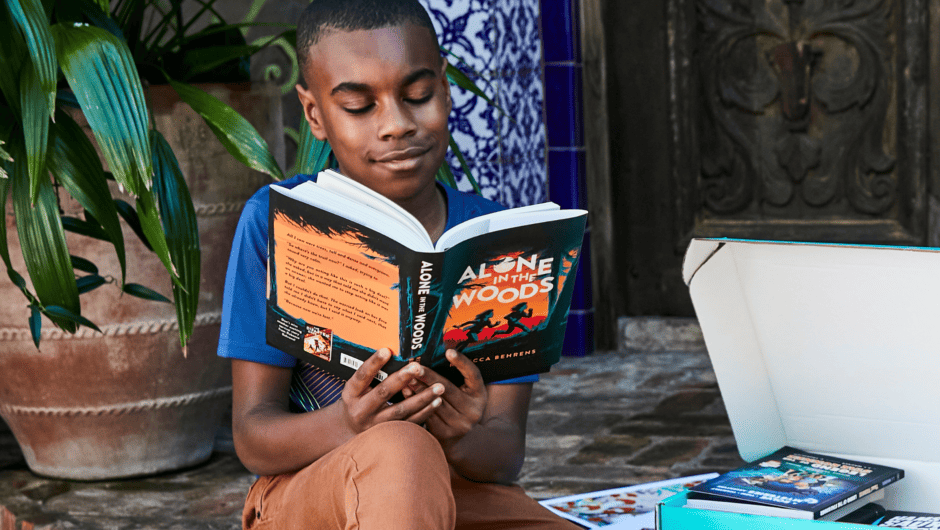 A boy is reading a novel while sitting near a Literati box on the stoop of his home.