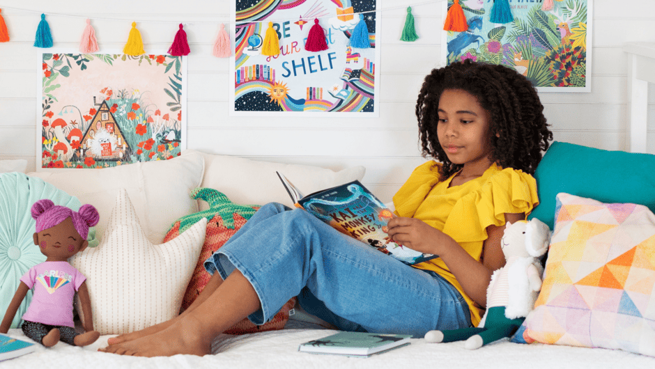 A girl reads a book while sitting on her bed full of colorful pillows and toys