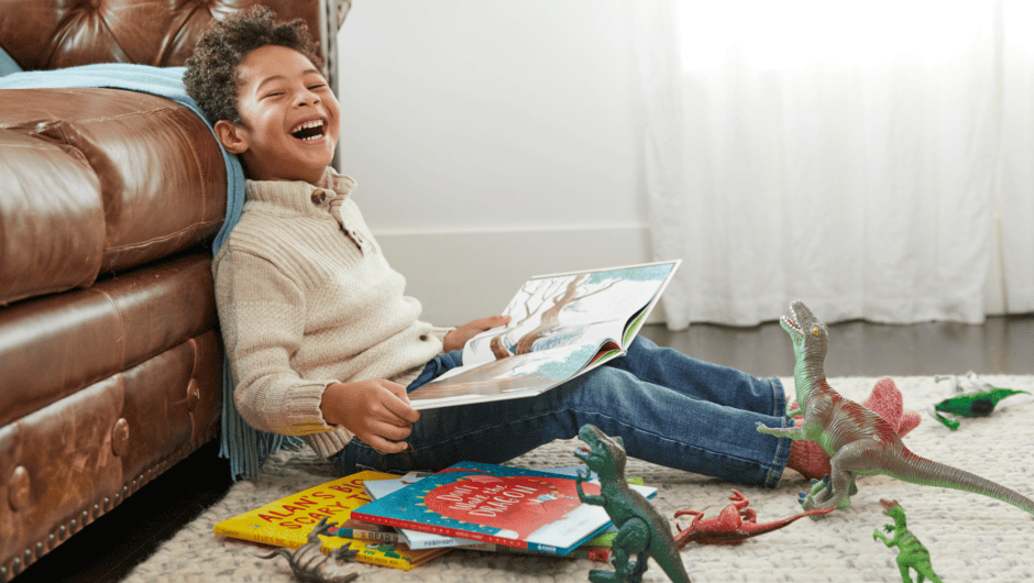 5 year old boy laughs as he reads a book with lots of toy dinosaurs on the floor.