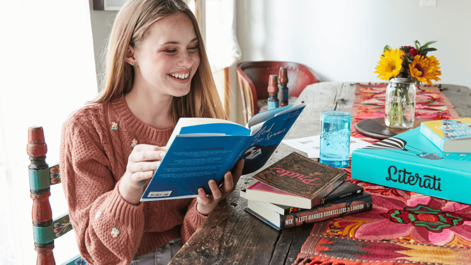 A teenage girl smiles as she reads a novel at the kitchen table. A Literati box is nearby.