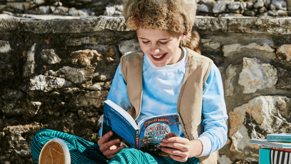 Young boy in fur hat sits against stone wall reading a book
