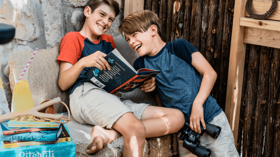Two boy laughing as they read a book in a fort. Literati box with books is on bench nearby.