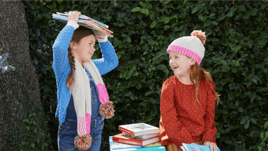 Two girls in fall clothes laugh in a garden with books and a Literati Box
