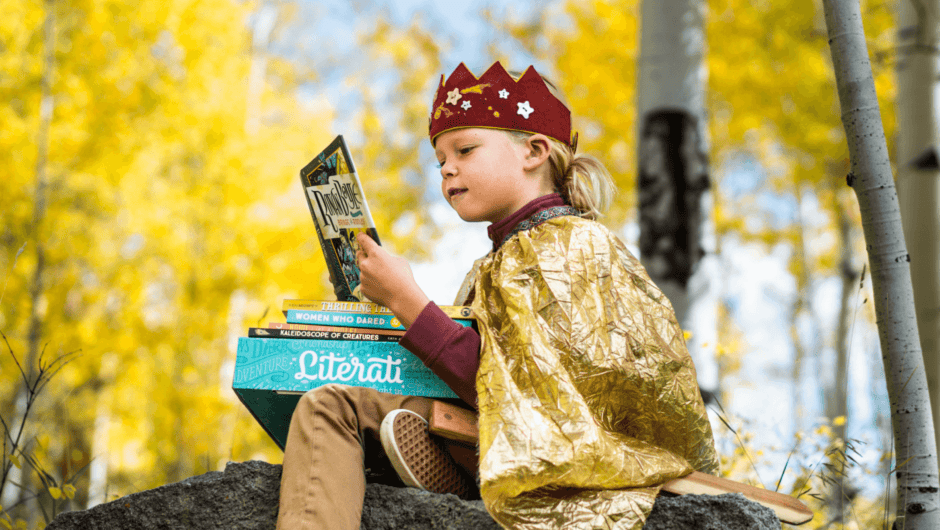 A young boy in a crown and cape reads a book in the woods.