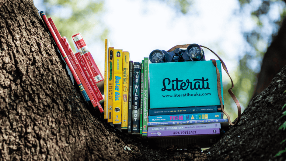 A colorful stack of books with a Literati box and binoculars sit in the nook of a tree.