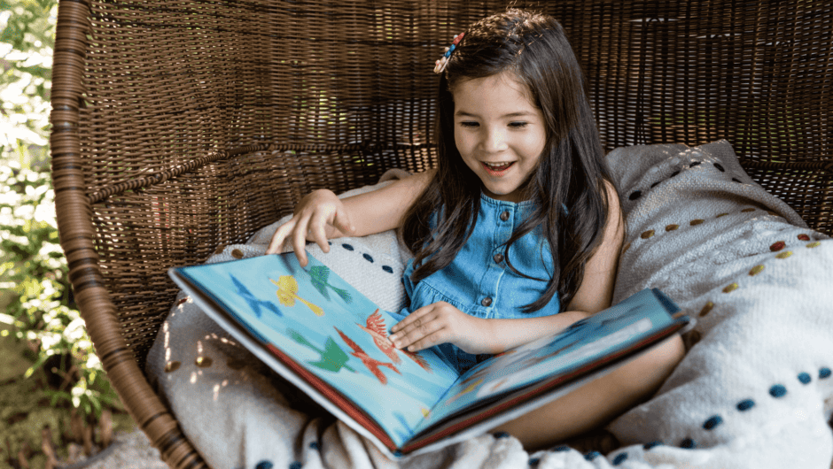 A young girl reads a picture book in a wicker chair