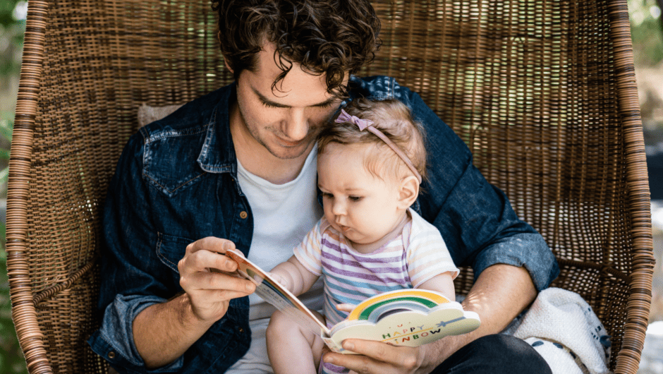 A father reads a rainbow book to a young baby girl in a wicker chair